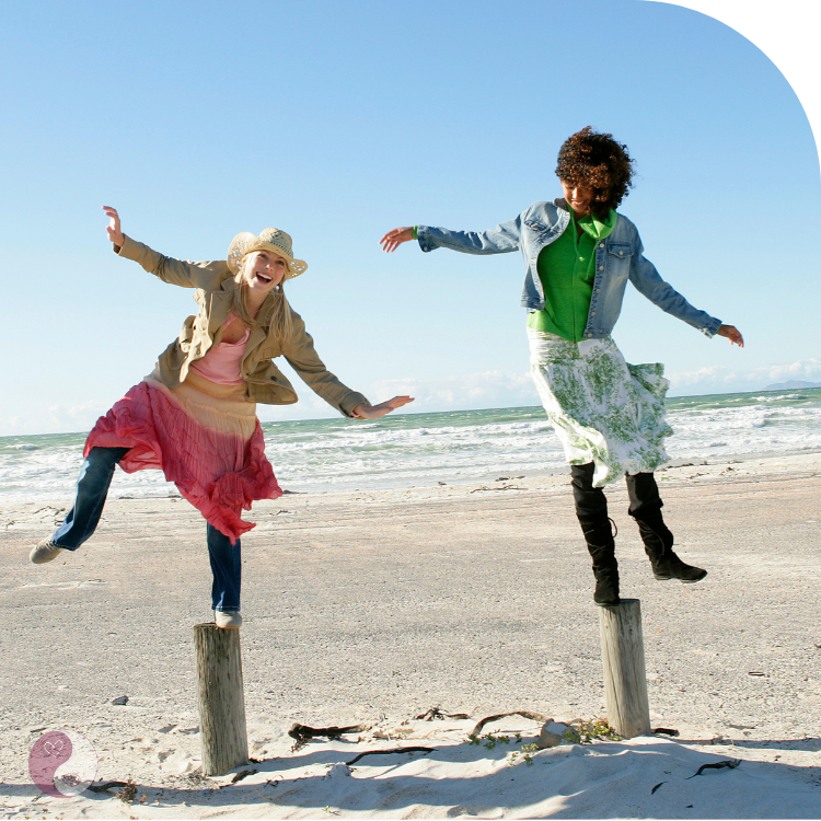 frauen-balancieren-holzpfaele-meer Zwei Frauen balancieren lachend auf Holzpfählen am Meer